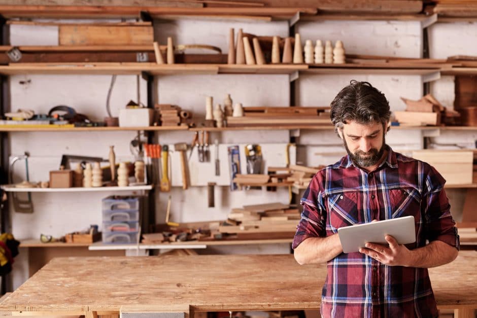 Artisan woodwork studio with shelving holding pieces of wood, with a carpenter standing in his workshop using a digital tablet