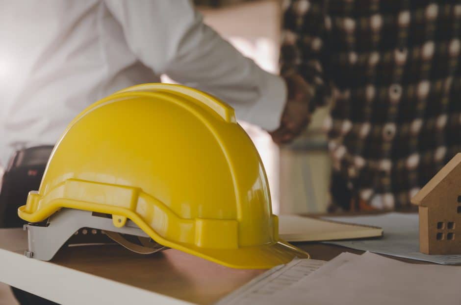 yellow safety helmet on workplace desk with construction worker team hands shaking